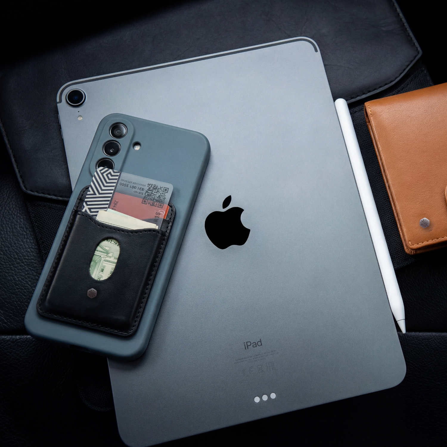 Smartphone with card holder on an iPad, next to a brown leather wallet on a dark surface